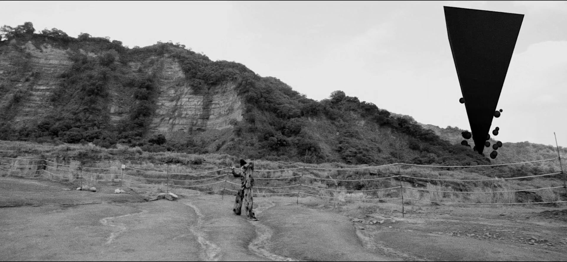 Monochrome rocky landscape with a figure and an inverted floating geometric form.